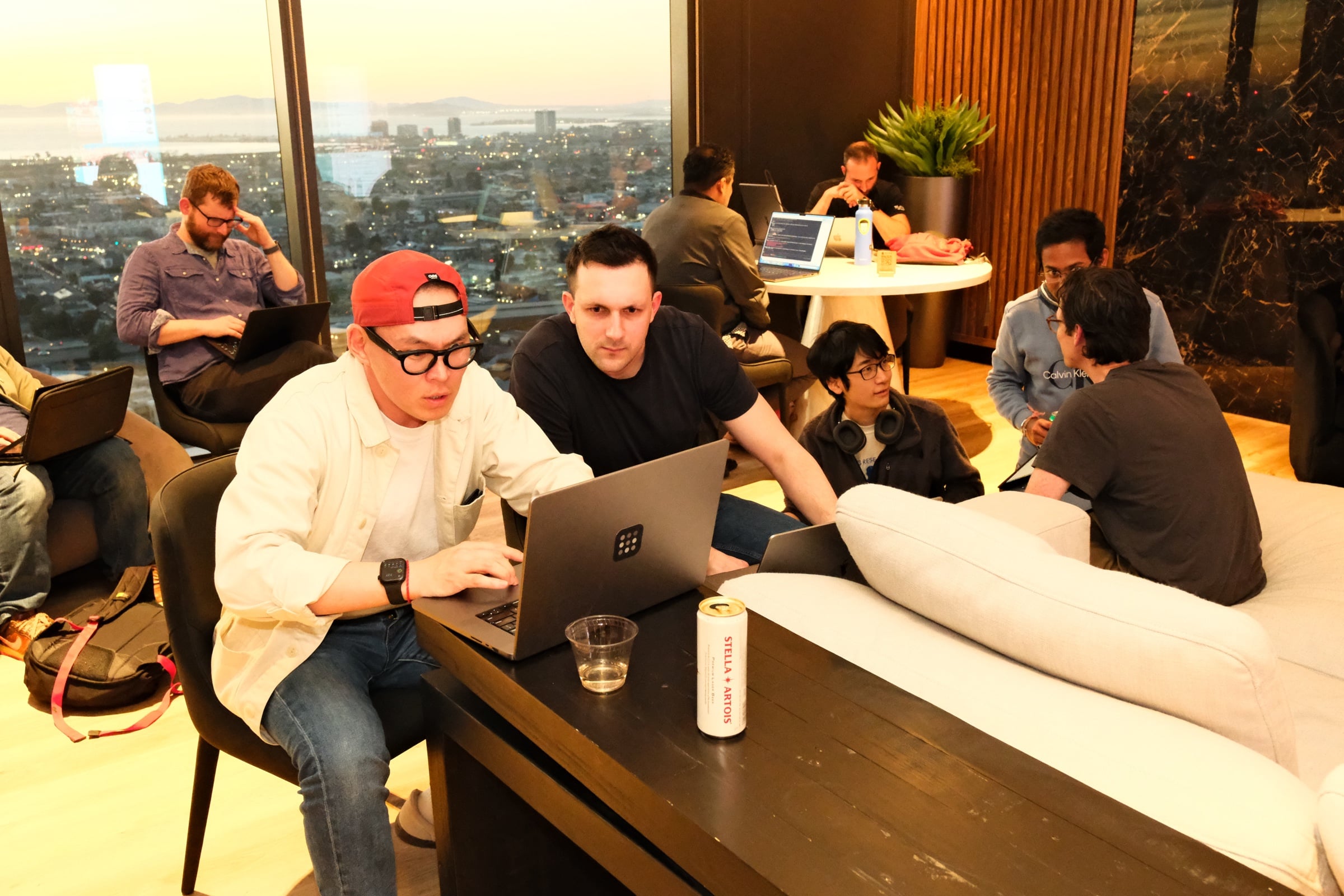 Two participants hacking at laptops, city lights through the penthouse windows behind them