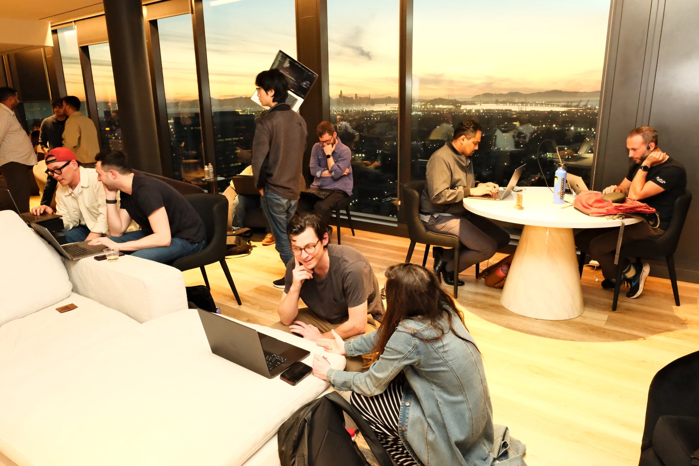 Participants hacking at laptops in the STAK penthouse, sunset through floor-to-ceiling windows with the Bay Area skyline
